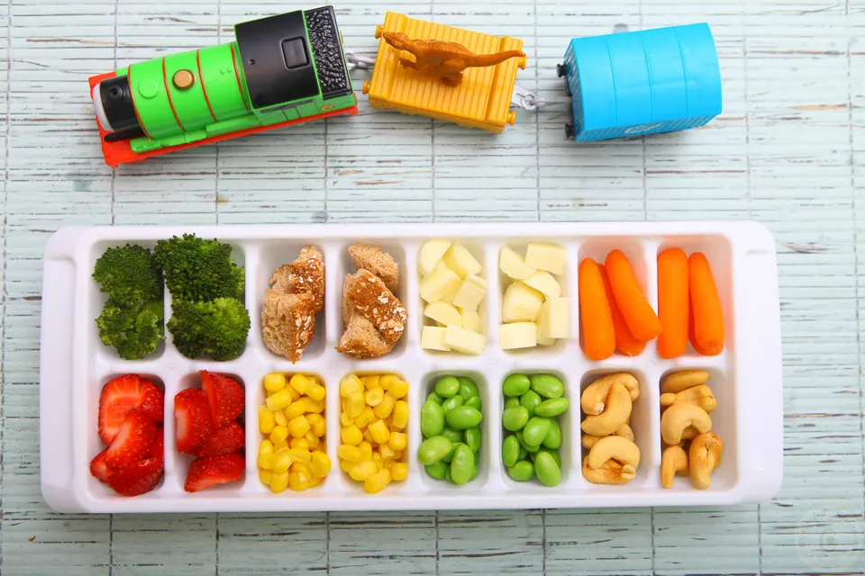 Ice cube tray filled with colorful toddler finger foods next to toy trains
